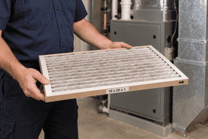 HVAC technician holding a dusty pleated furnace filter with its size label visible.